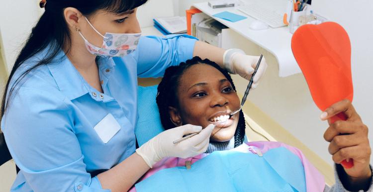 A dentist who is a woman behind the patient with dental tools in her hands. The patient has a blue sheet protecting her clothes, she is also opening her mouth out wide for her treatment and looking into a mirror.