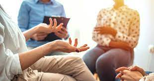 A group of three people sitting in a circle, a person on the left is wearing brown trousers and talking using hand gestures. A person in the middle is wearing a smart blue button-up shirt and is holding a clipboard to record down notes. Also a person at the right listening.