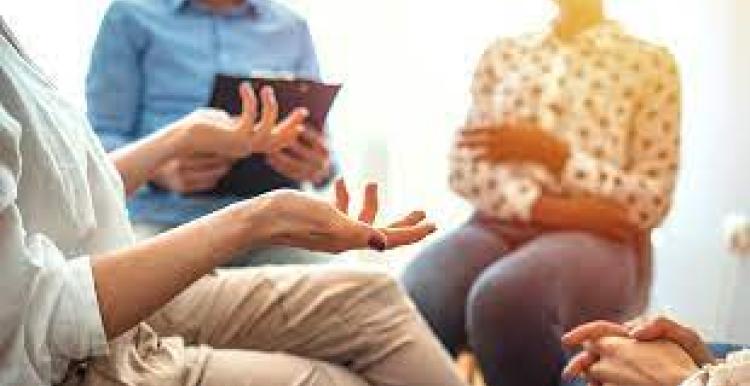 A group of three people sitting in a circle, a person on the left is wearing brown trousers and talking using hand gestures. A person in the middle is wearing a smart blue button-up shirt and is holding a clipboard to record down notes. Also a person at the right listening.