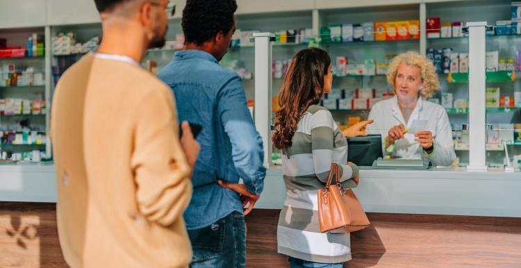 This is a scene of a pharmacy. The pharmacist has white skin, short curly blonde hair and is wearing a white lab coat. She is behind a white counter. There are three patients in the queue in front of the counter. The first patient is a woman with brown curly hair, checkered coat and is holding a handbag on her arm. The man behind her has black hair and is wearing a denim outfit. The final patient is a bearded man with brown hair and wearing a yellow jumper.