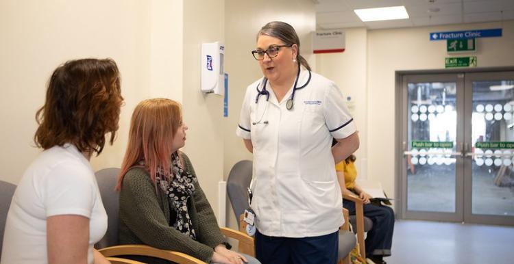 This is a hospital corridor. Two women are sitting on chairs on the left hand side. One woman is brown haired, tanned and is wearing a white unsleeved t-shirt. The second woman is wearing a blouse, cardigan and has ginger hair. There is a female health and social care professional speaking to the two women. This professional is brown haired, wearing glasses and wearing a white scrub uniform.