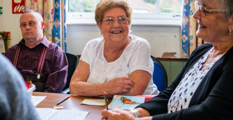 There are three people sitting on a table. There is a man on the left wearing a checkered shirt with suspenders. There is a lady in the middle wearing glasses and a white half-sleeved top, she is also smiling to the right. At the right is another lady wearing glasses, a black cardigan and white top.