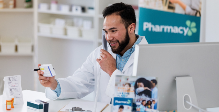 There is a male pharmacist who has dark brown hair and a beard. He is wearing a blue button up shirt with a lab coat on top of that. He is holding a white packet of tablets. On the right hand side there is a Macbook.