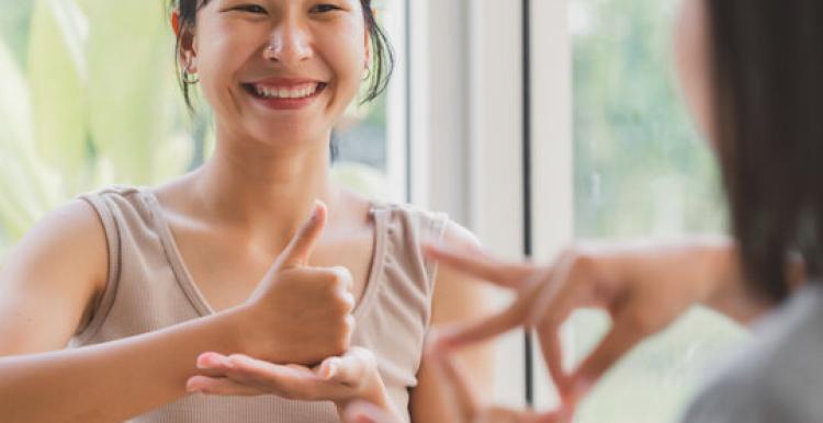 There is one lady in this picture. On the right hand side corner, we can see her shoulder and left side of her hair as she is facing a mirror. In the middle of the photo, there is a woman who has black hair with a fringe and she is smiling while practicing British Sign Language