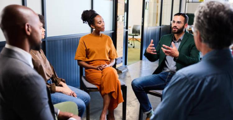 This photo is showing a focus group and there are four people. There is one man on the left hand side and one man on the right hand side, both of them are facing two other people in front. So the faces of these men cannot be seen. In the middle of the photo is a lady wearing an orange dress and a man sitting beside her, talking to the group. The men are wearing smart suits.