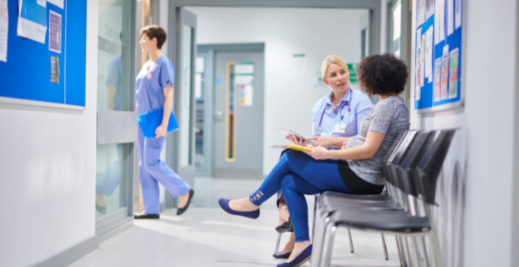 This is a picture of an NHS facility. There is a blue notice board with a white frame and there are some staff pictures posted on there. On the right hand side there is a line of chairs. There is a brown skinned woman with curly Afro hair and she is speaking to a white, blonde haired woman.