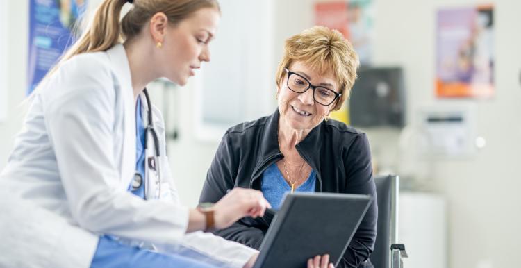 On the left hand side there is a female doctor. She is wearing blue scrubs, white lab coat and has a stethoscope. In her hand is a tablet to show a patient some information. In the middle is a patient. She is wearing a blue top, black jacket and she wears red glasses. She has short blonde hair and is smiling while looking at the tablet.