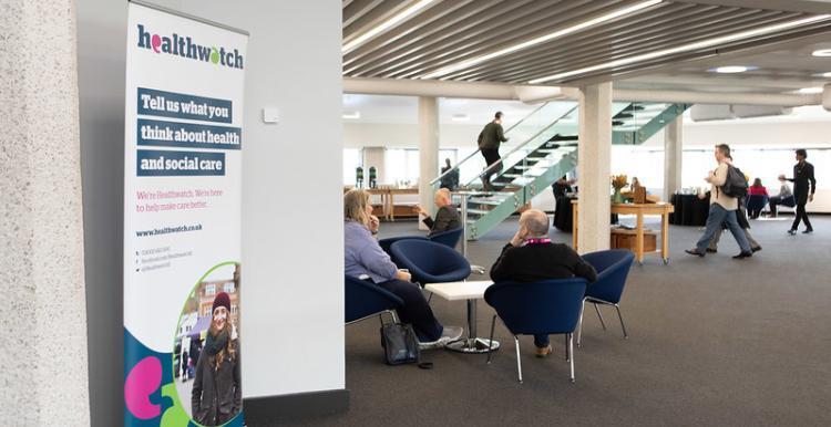 This photo is showing a seminar building. On the left hand side there is a plain white wall and infront of the wall is a Healthwatch pull-up sign. In the middle of the room there is a group of people sitting on blue comfy chairs around a white table.
