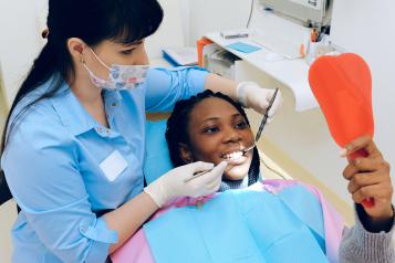 A dentist who is a woman behind the patient with dental tools in her hands. The patient has a blue sheet protecting her clothes, she is also opening her mouth out wide for her treatment and looking into a mirror.