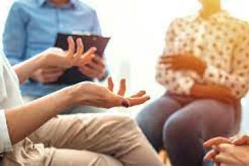 A group of three people sitting in a circle, a person on the left is wearing brown trousers and talking using hand gestures. A person in the middle is wearing a smart blue button-up shirt and is holding a clipboard to record down notes. Also a person at the right listening.