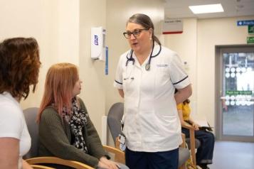 This is a hospital corridor. Two women are sitting on chairs on the left hand side. One woman is brown haired, tanned and is wearing a white unsleeved t-shirt. The second woman is wearing a blouse, cardigan and has ginger hair. There is a female health and social care professional speaking to the two women. This professional is brown haired, wearing glasses and wearing a white scrub uniform.