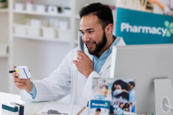 There is a male pharmacist who has dark brown hair and a beard. He is wearing a blue button up shirt with a lab coat on top of that. He is holding a white packet of tablets. On the right hand side there is a Macbook.