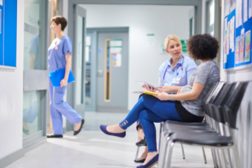 This is a picture of an NHS facility. There is a blue notice board with a white frame and there are some staff pictures posted on there. On the right hand side there is a line of chairs. There is a brown skinned woman with curly Afro hair and she is speaking to a white, blonde haired woman.