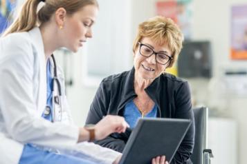 On the left hand side there is a female doctor. She is wearing blue scrubs, white lab coat and has a stethoscope. In her hand is a tablet to show a patient some information. In the middle is a patient. She is wearing a blue top, black jacket and she wears red glasses. She has short blonde hair and is smiling while looking at the tablet.