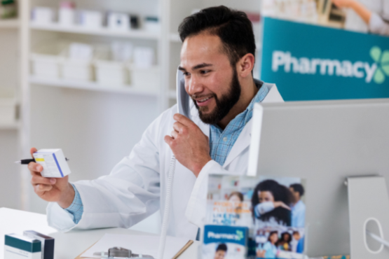 There is a male pharmacist who has dark brown hair and a beard. He is wearing a blue button up shirt with a lab coat on top of that. He is holding a white packet of tablets. On the right hand side there is a Macbook.