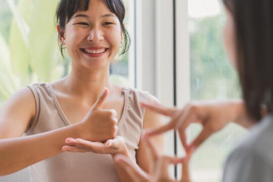 There is one lady in this picture. On the right hand side corner, we can see her shoulder and left side of her hair as she is facing a mirror. In the middle of the photo, there is a woman who has black hair with a fringe and she is smiling while practicing British Sign Language
