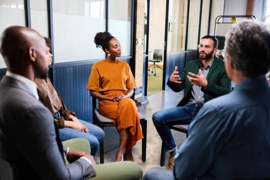 This photo is showing a focus group and there are four people. There is one man on the left hand side and one man on the right hand side, both of them are facing two other people in front. So the faces of these men cannot be seen. In the middle of the photo is a lady wearing an orange dress and a man sitting beside her, talking to the group. The men are wearing smart suits.