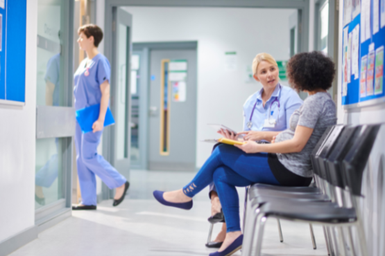This is a picture of an NHS facility. There is a blue notice board with a white frame and there are some staff pictures posted on there. On the right hand side there is a line of chairs. There is a brown skinned woman with curly Afro hair and she is speaking to a white, blonde haired woman.