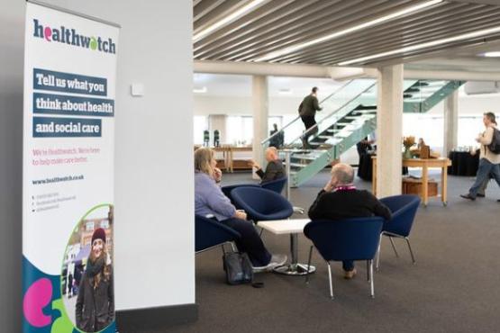 This photo is showing a seminar building. On the left hand side there is a plain white wall and infront of the wall is a Healthwatch pull-up sign. In the middle of the room there is a group of people sitting on blue comfy chairs around a white table.