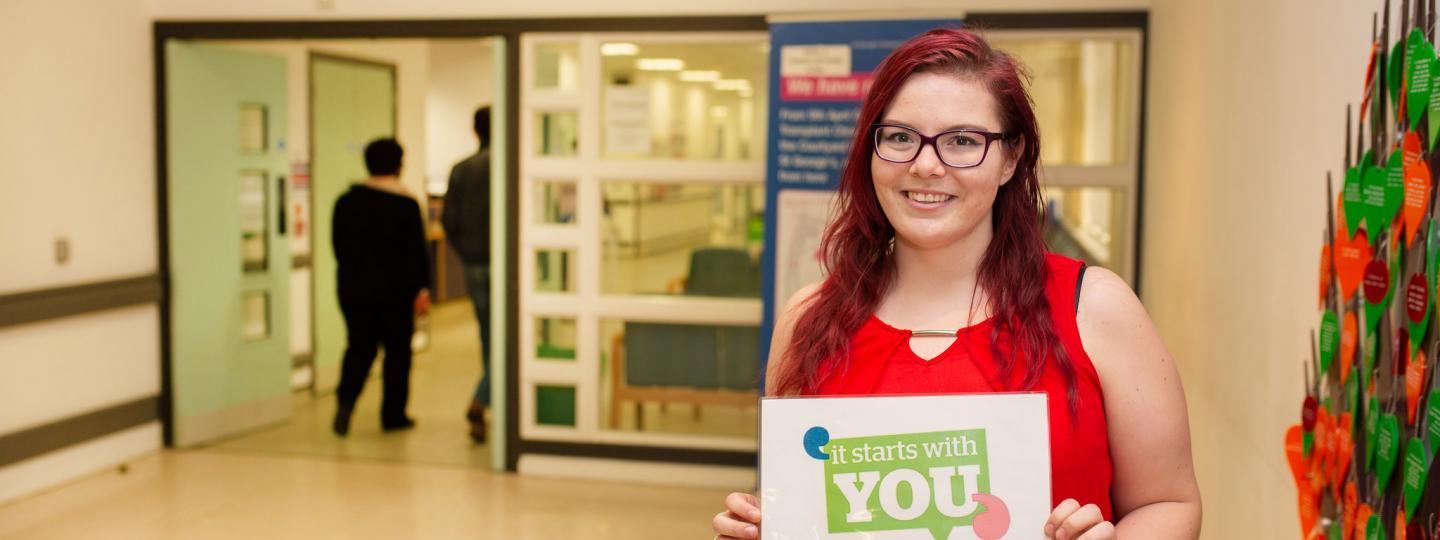 Woman stood in a hospital corridor holding a sign "It starts with you"
