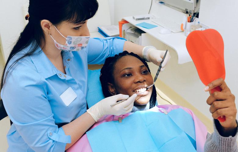 A dentist who is a woman behind the patient with dental tools in her hands. The patient has a blue sheet protecting her clothes, she is also opening her mouth out wide for her treatment and looking into a mirror.