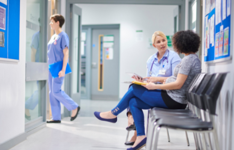 This is a picture of an NHS facility. There is a blue notice board with a white frame and there are some staff pictures posted on there. On the right hand side there is a line of chairs. There is a brown skinned woman with curly Afro hair and she is speaking to a white, blonde haired woman.