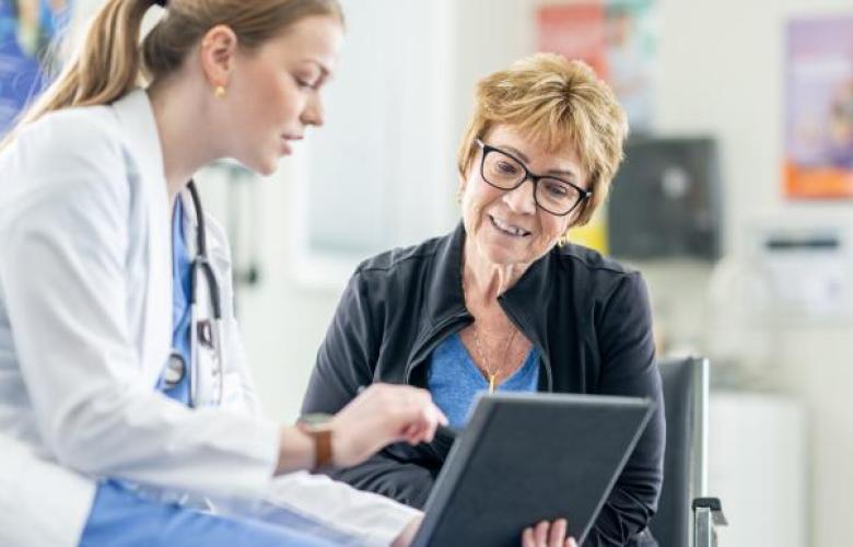 On the left hand side there is a female doctor. She is wearing blue scrubs, white lab coat and has a stethoscope. In her hand is a tablet to show a patient some information. In the middle is a patient. She is wearing a blue top, black jacket and she wears red glasses. She has short blonde hair and is smiling while looking at the tablet.