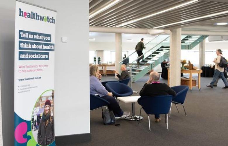 This photo is showing a seminar building. On the left hand side there is a plain white wall and infront of the wall is a Healthwatch pull-up sign. In the middle of the room there is a group of people sitting on blue comfy chairs around a white table.
