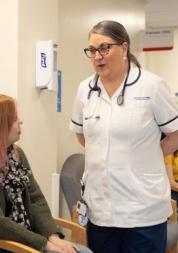 This is a hospital corridor. Two women are sitting on chairs on the left hand side. One woman is brown haired, tanned and is wearing a white unsleeved t-shirt. The second woman is wearing a blouse, cardigan and has ginger hair. There is a female health and social care professional speaking to the two women. This professional is brown haired, wearing glasses and wearing a white scrub uniform.
